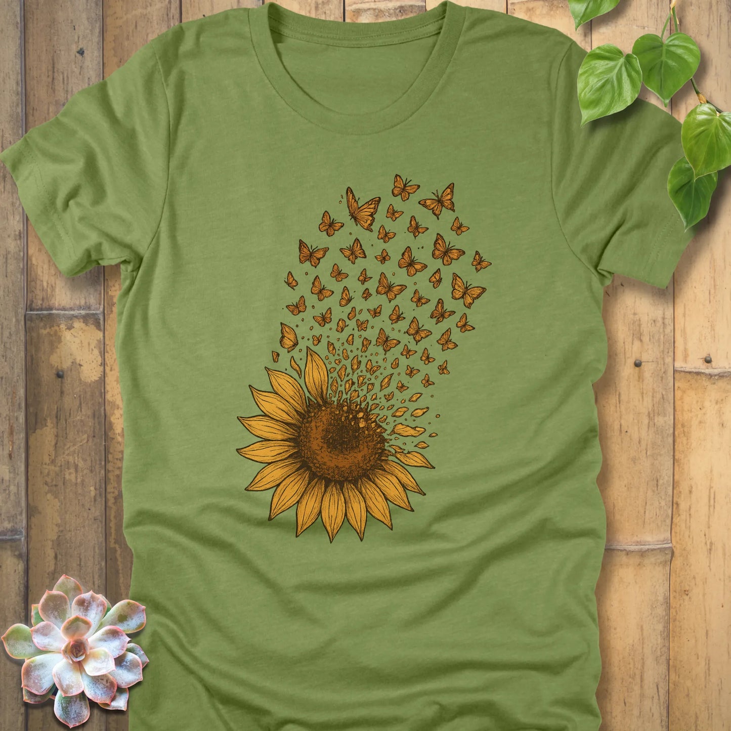 Heather Green t-shirt showcasing a bright sunflower graphic, petals scattering upward as butterflies