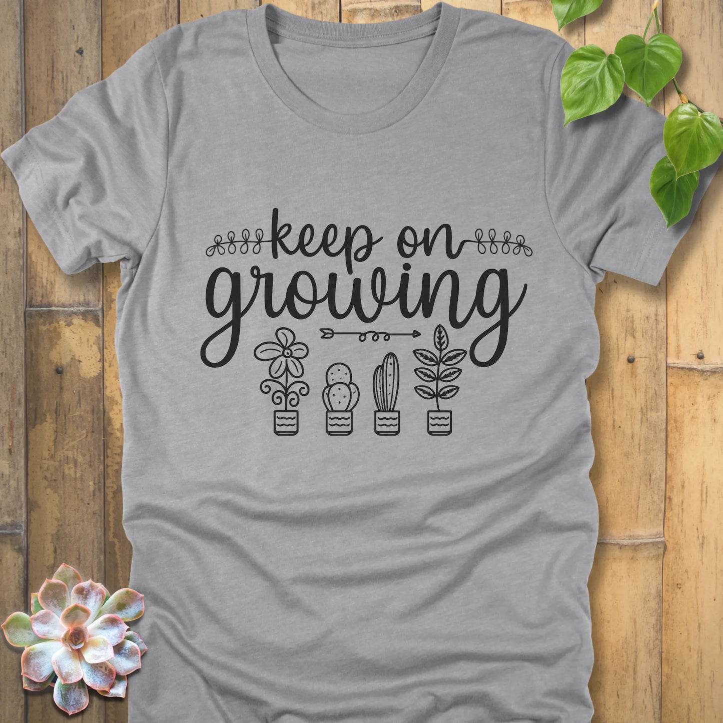Athletic Heather t-shirt with 'Keep on Growing' text in cursive above four potted plant illustrations, featuring flower, cactus, succulent, and leafy plant designs. Garden-themed graphic tee.