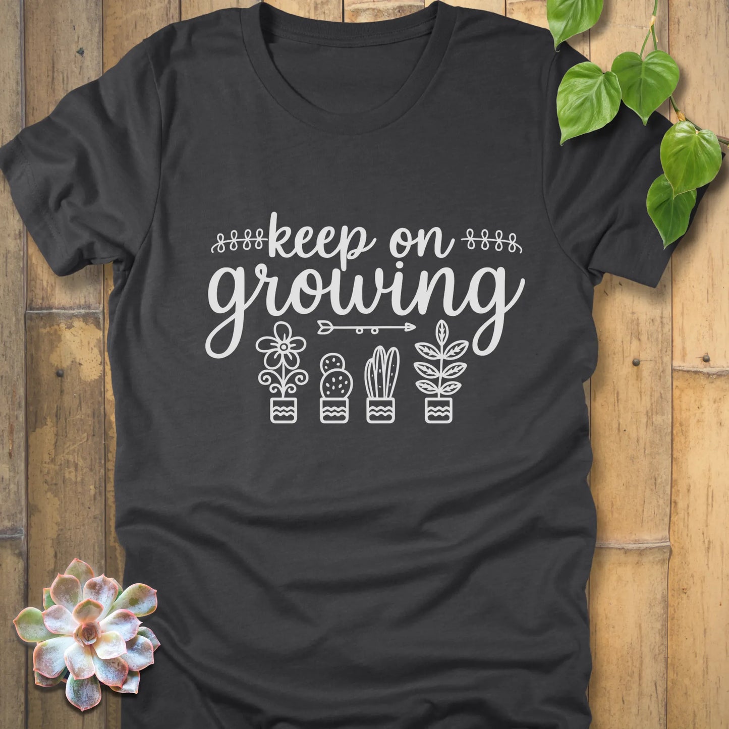 Dark Grey Heather t-shirt with 'Keep on Growing' text in cursive above four potted plant illustrations, featuring flower, cactus, succulent, and leafy plant designs. Garden-themed graphic tee.