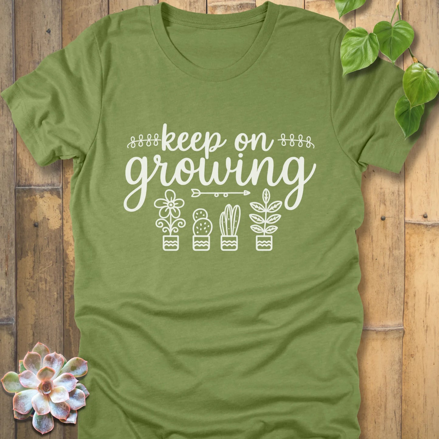 Heather Green t-shirt with 'Keep on Growing' text in cursive above four potted plant illustrations, featuring flower, cactus, succulent, and leafy plant designs. Garden-themed graphic tee.