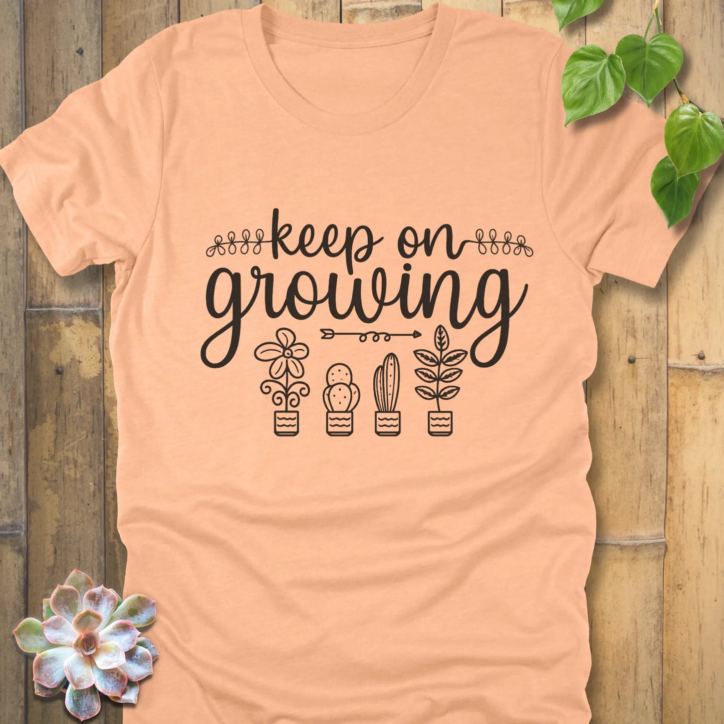 Heather Peach t-shirt with 'Keep on Growing' text in cursive above four potted plant illustrations, featuring flower, cactus, succulent, and leafy plant designs. Garden-themed graphic tee.