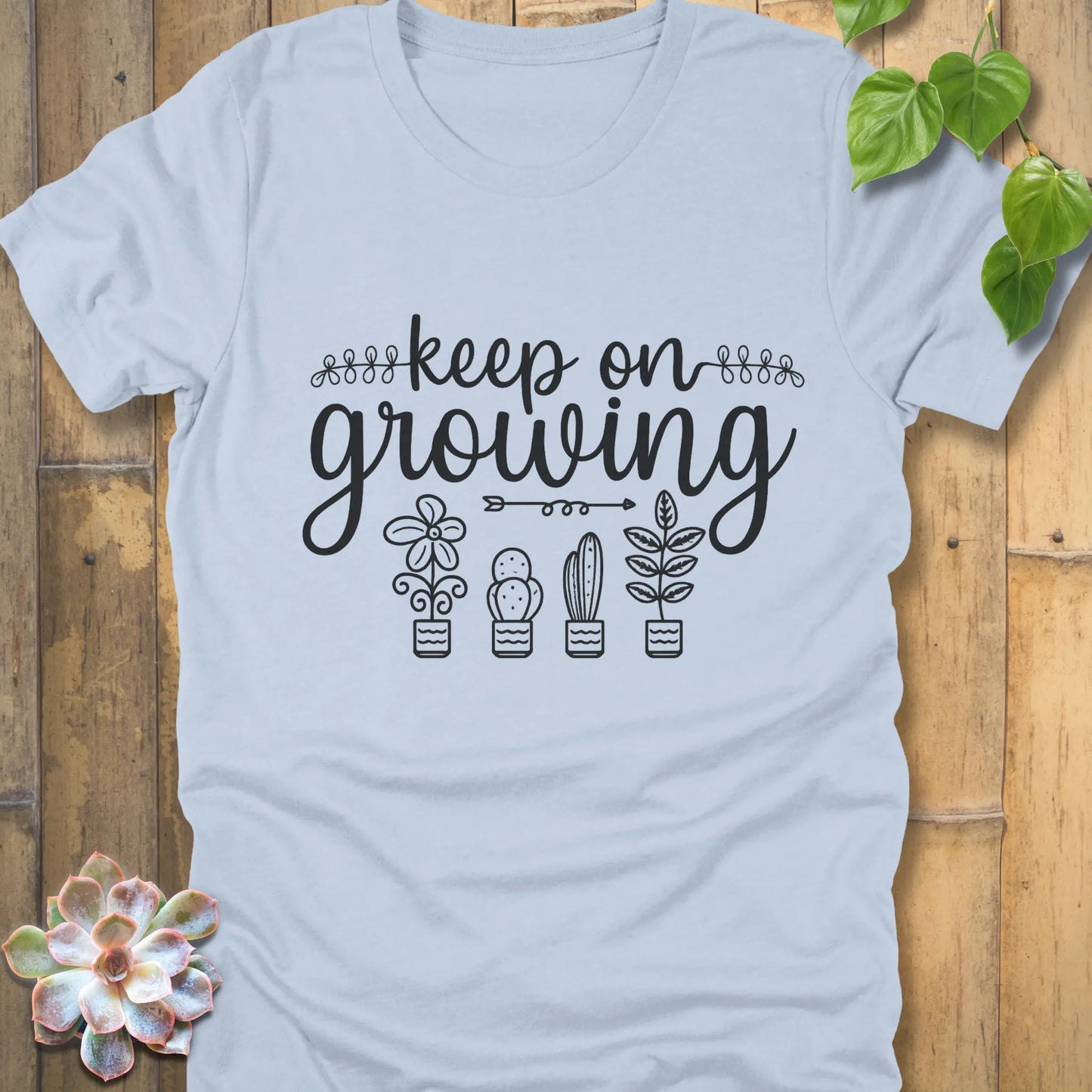 Light Blue t-shirt with 'Keep on Growing' text in cursive above four potted plant illustrations, featuring flower, cactus, succulent, and leafy plant designs. Garden-themed graphic tee.