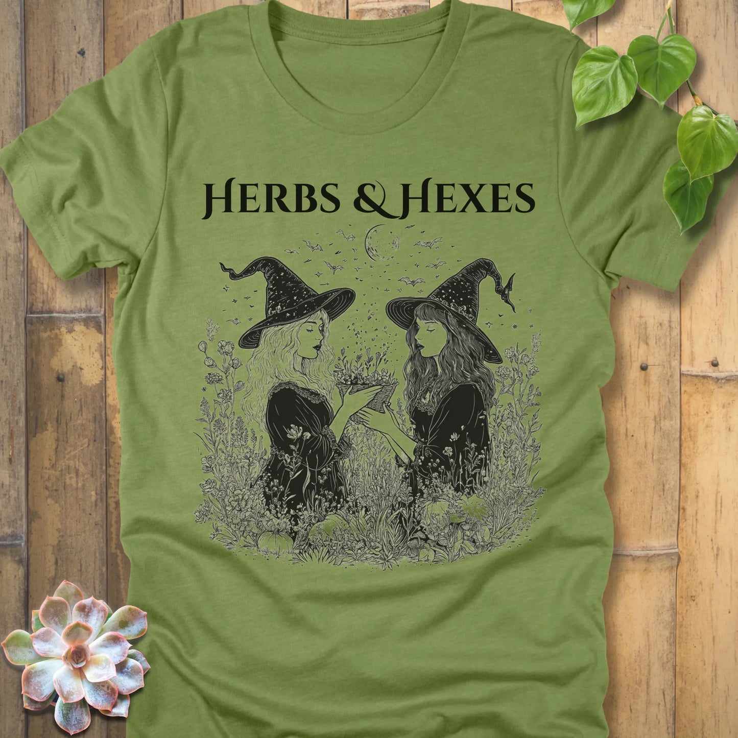 Heather Green t-shirt featuring the design Herbs & Hexes in black letters, printed with two witches