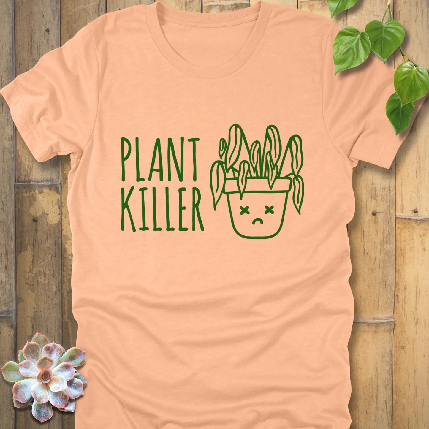 Heather peach t-shirt featuring the design Plant Killer printed in green letters with a plant in an emoji pot