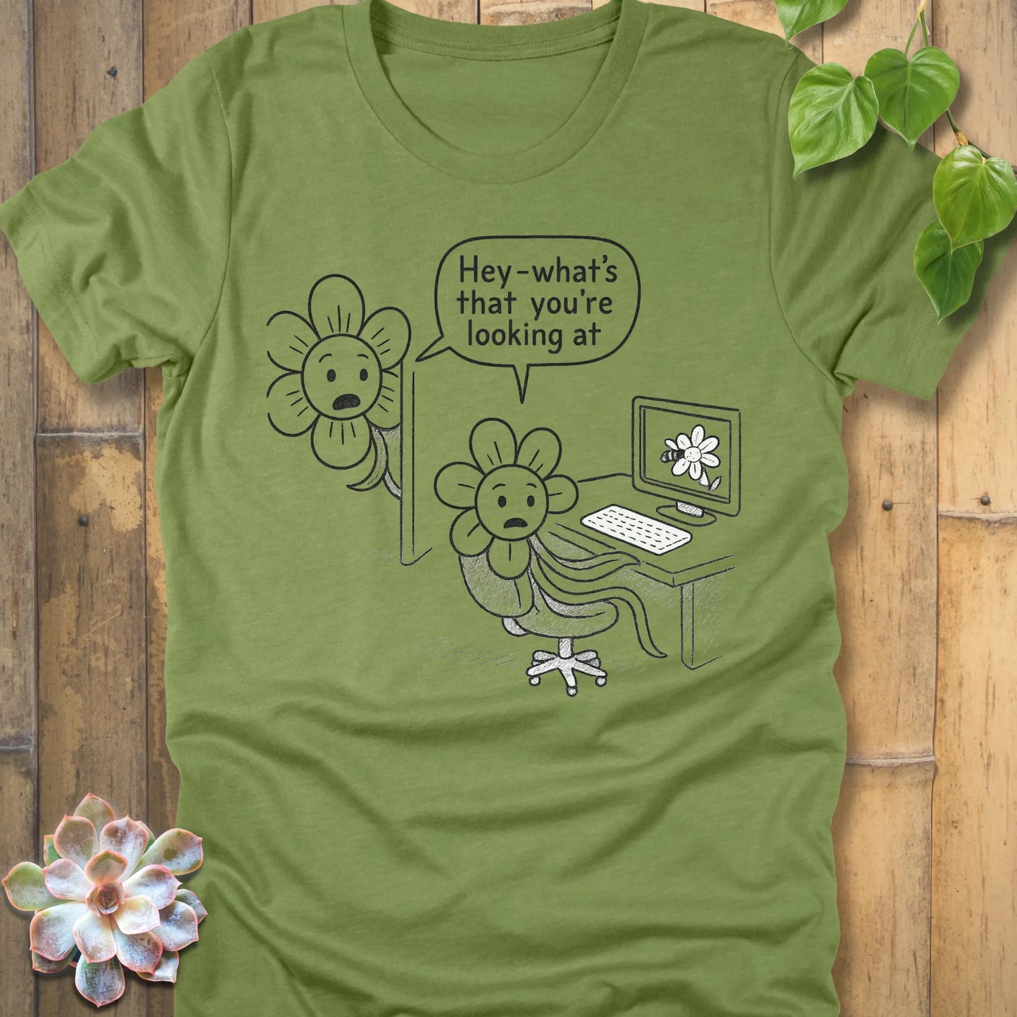 Heather green unisex Bella+Canvas 3001 T-shirt featuring a cartoon flower sitting at a desk, caught looking at a computer screen with a flower image. Another flower leans around the monitor and says in a speech bubble, ‘Hey—what’s that you’re looking at?