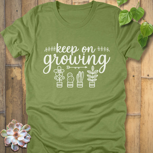 Heather Green t-shirt with 'Keep on Growing' text in cursive above four potted plant illustrations, featuring flower, cactus, succulent, and leafy plant designs. Garden-themed graphic tee.