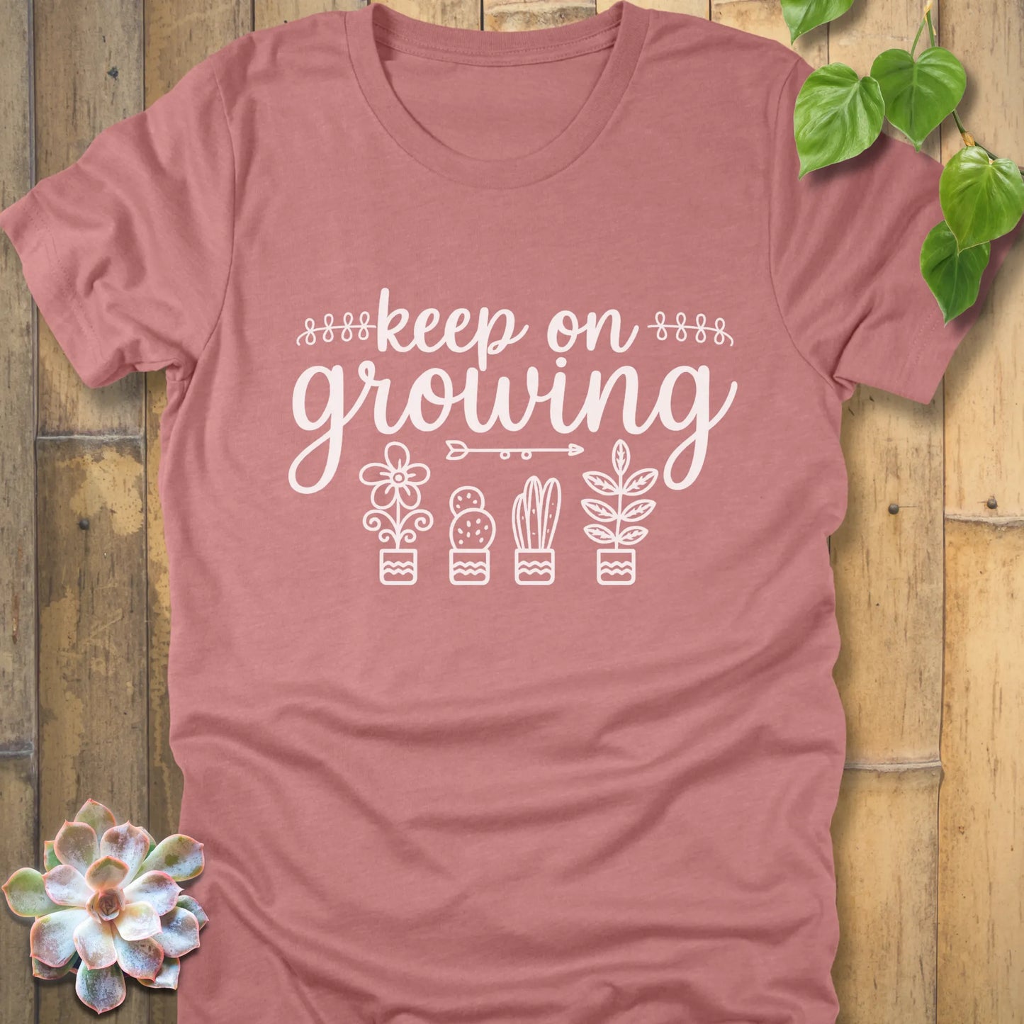 Heather Mauve t-shirt with 'Keep on Growing' text in cursive above four potted plant illustrations, featuring flower, cactus, succulent, and leafy plant designs. Garden-themed graphic tee.