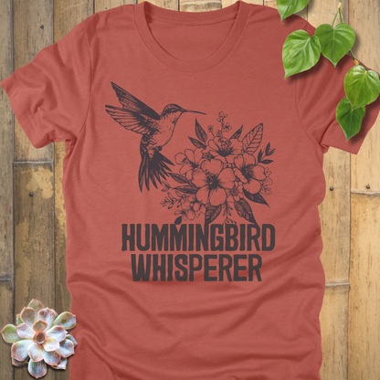 Heather clay “Hummingbird Whisperer” T-shirt featuring a detailed black illustration of a hummingbird hovering near blooming flowers, printed on soft Bella Canvas fabric