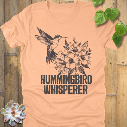 Heather peach “Hummingbird Whisperer” graphic tee showing a hummingbird and floral bouquet in black ink, lightweight comfortable shirt for bird lovers