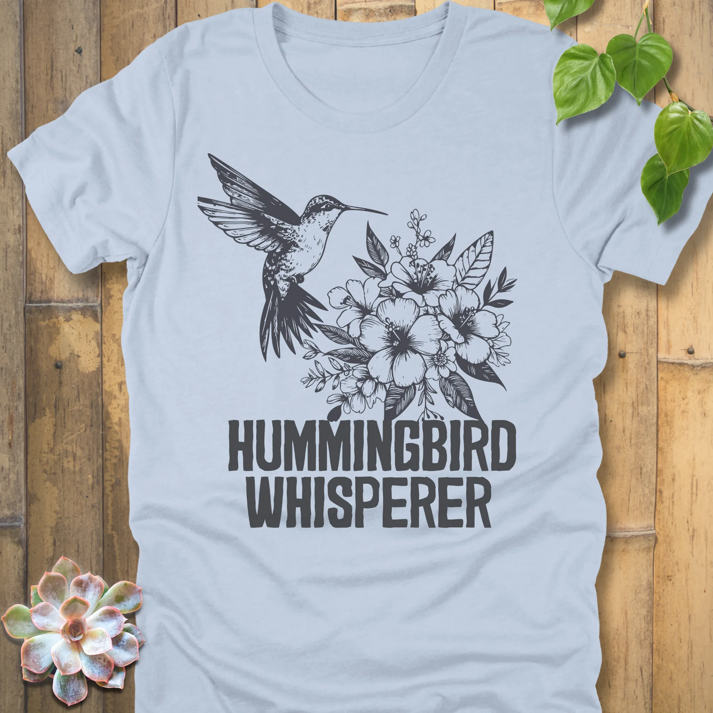 Light blue “Hummingbird Whisperer” T-shirt featuring a delicate black hummingbird hovering beside blossoms, soft breathable unisex tee.