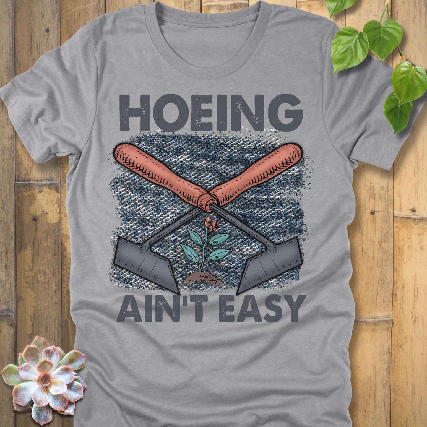 Athletic Heather t-shirt featuring the design Hoeing Ain’t Easy with gardening tools and plants printed in bold letters, funny gardening shirt.