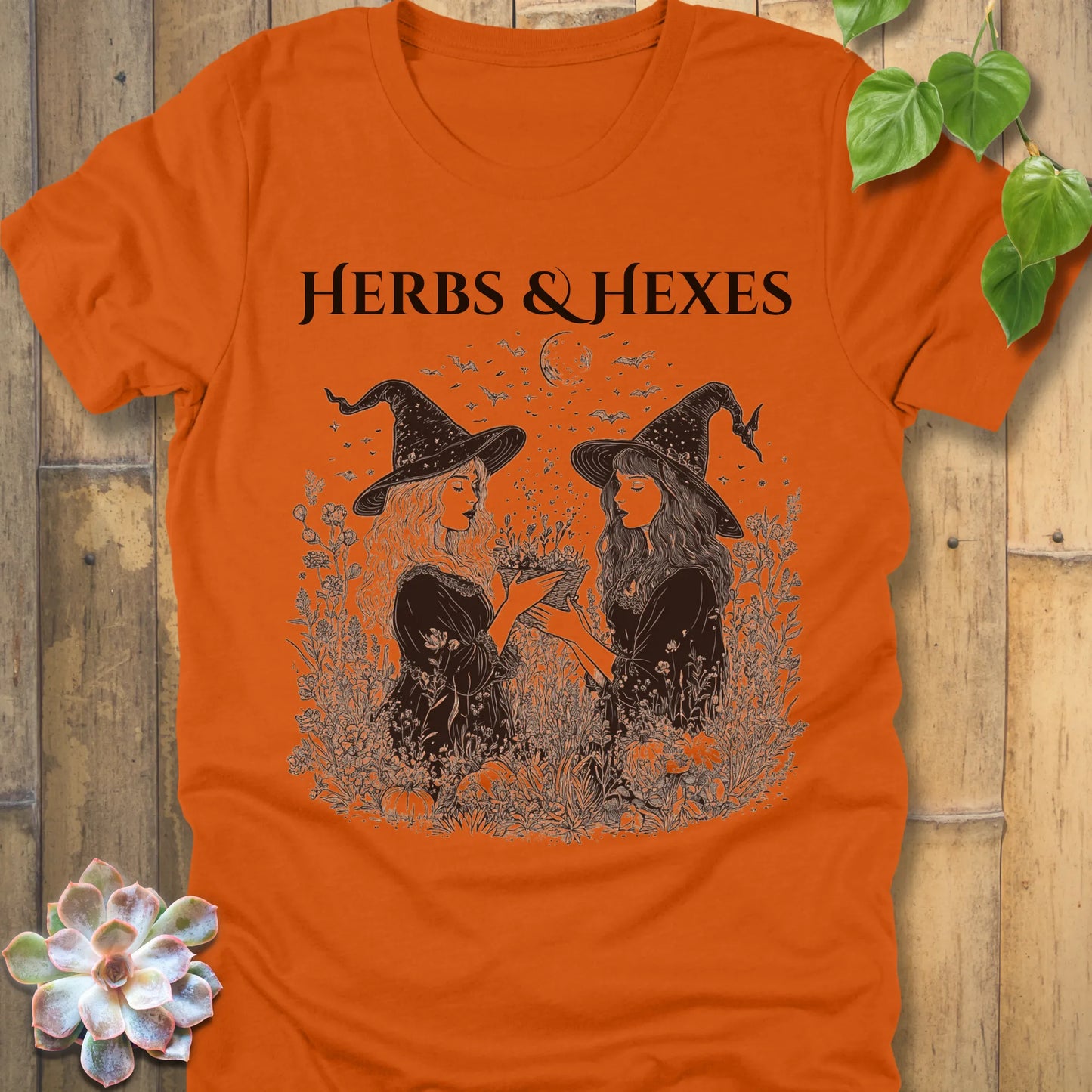 Autumn t-shirt featuring the design Herbs & Hexes in black letters, printed with two witches