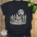 A black short-sleeve graphic t-shirt laid flat on a rustic wooden background, featuring a detailed black and white illustration of a rooster, two hens, a picket fence, and a full moon. Decorative green leaves and a succulent are placed around the shirt.