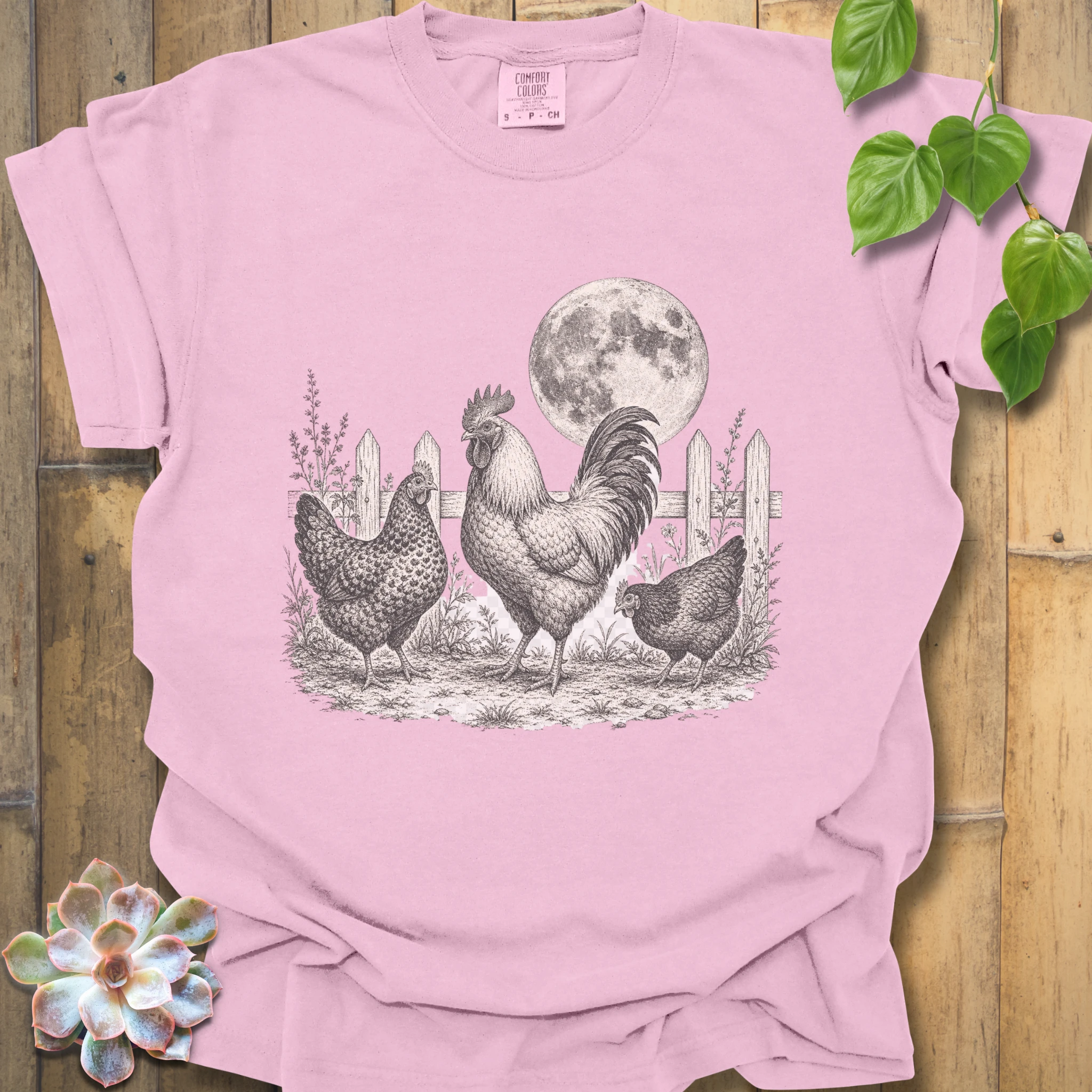 A light pink short-sleeve t-shirt with a detailed black and white graphic of three chickens and a rooster in front of a full moon and picket fence. The shirt is displayed in a flat-lay arrangement on a wooden background with decorative green leaves and a succulent.