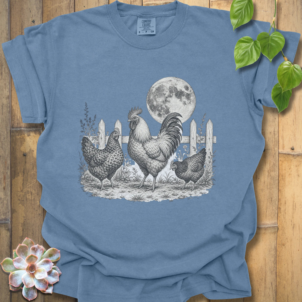 A blue short-sleeve t-shirt with a detailed black and white graphic of a rooster, hen, chick, and a full moon behind a picket fence, displayed on a wooden background with decorative green leaves and a succulent.