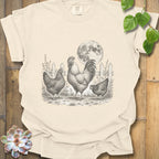 A cream-colored short-sleeve t-shirt with a black and white graphic of a rooster, two hens, and a full moon, laid flat on a rustic wooden background with decorative green leaves and a succulent.