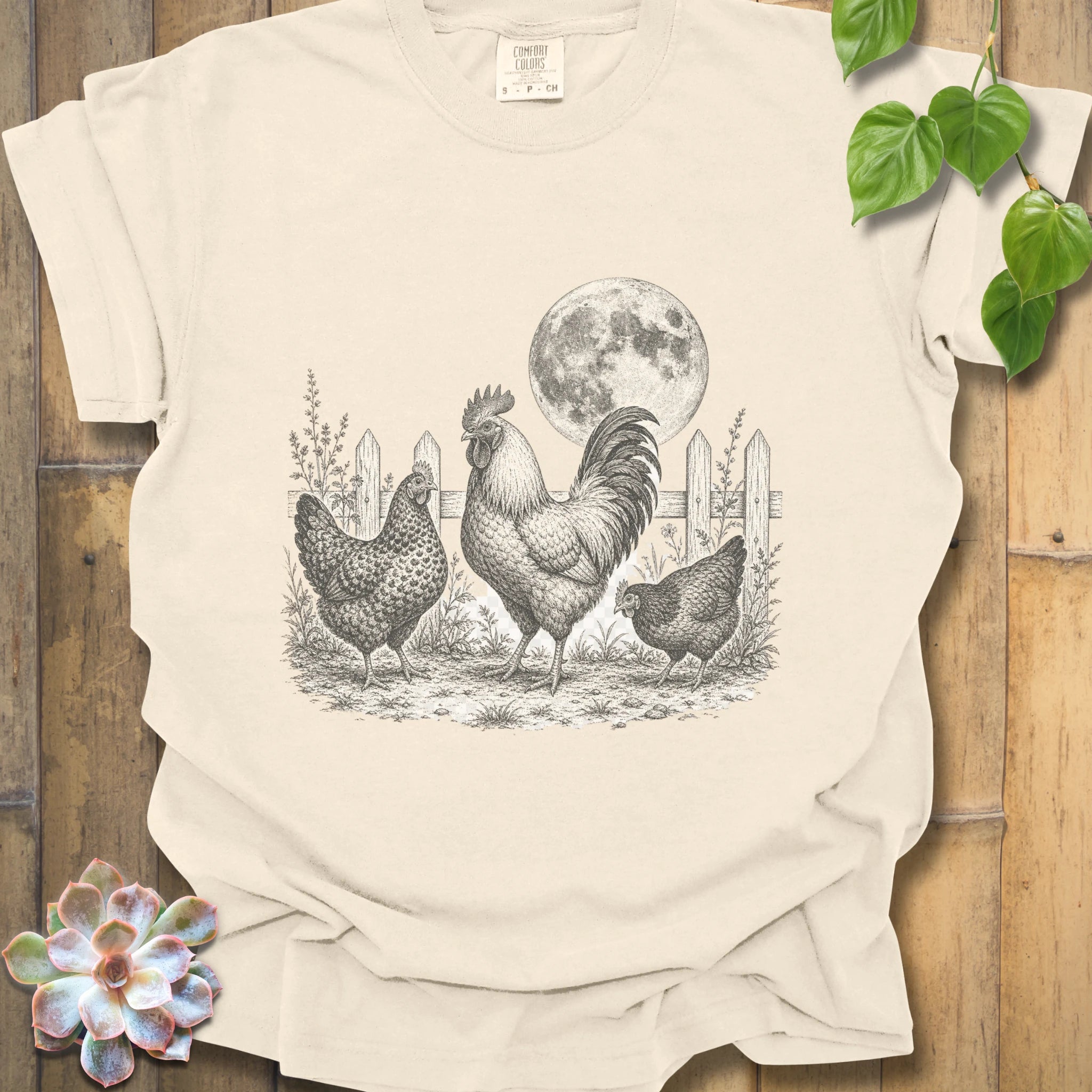 A cream-colored short-sleeve t-shirt with a black and white graphic of a rooster, two hens, and a full moon, laid flat on a rustic wooden background with decorative green leaves and a succulent.