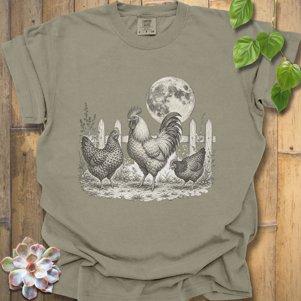 An olive green short-sleeve t-shirt with a detailed black and white graphic of a rooster, two hens, and a full moon in a barn setting, displayed on a rustic wooden background with decorative green leaves and a succulent.