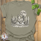 An olive green short-sleeve t-shirt with a detailed black and white graphic of a rooster, two hens, and a full moon in a barn setting, displayed on a rustic wooden background with decorative green leaves and a succulent.
