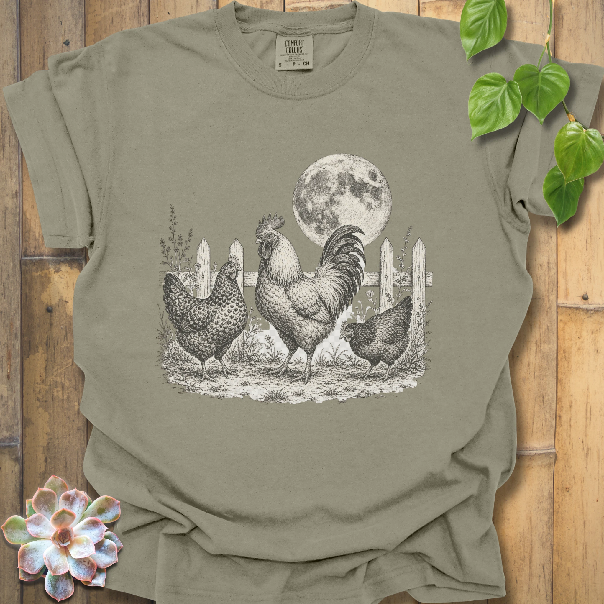An olive green short-sleeve t-shirt with a detailed black and white graphic of a rooster, two hens, and a full moon in a barn setting, displayed on a rustic wooden background with decorative green leaves and a succulent.