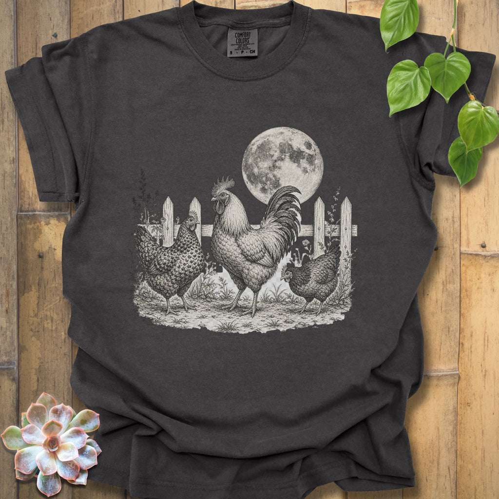 A dark charcoal gray t-shirt with a detailed black and white graphic of a rooster, two hens, a picket fence, and a full moon, displayed on a wooden background with decorative green leaves and a succulent.