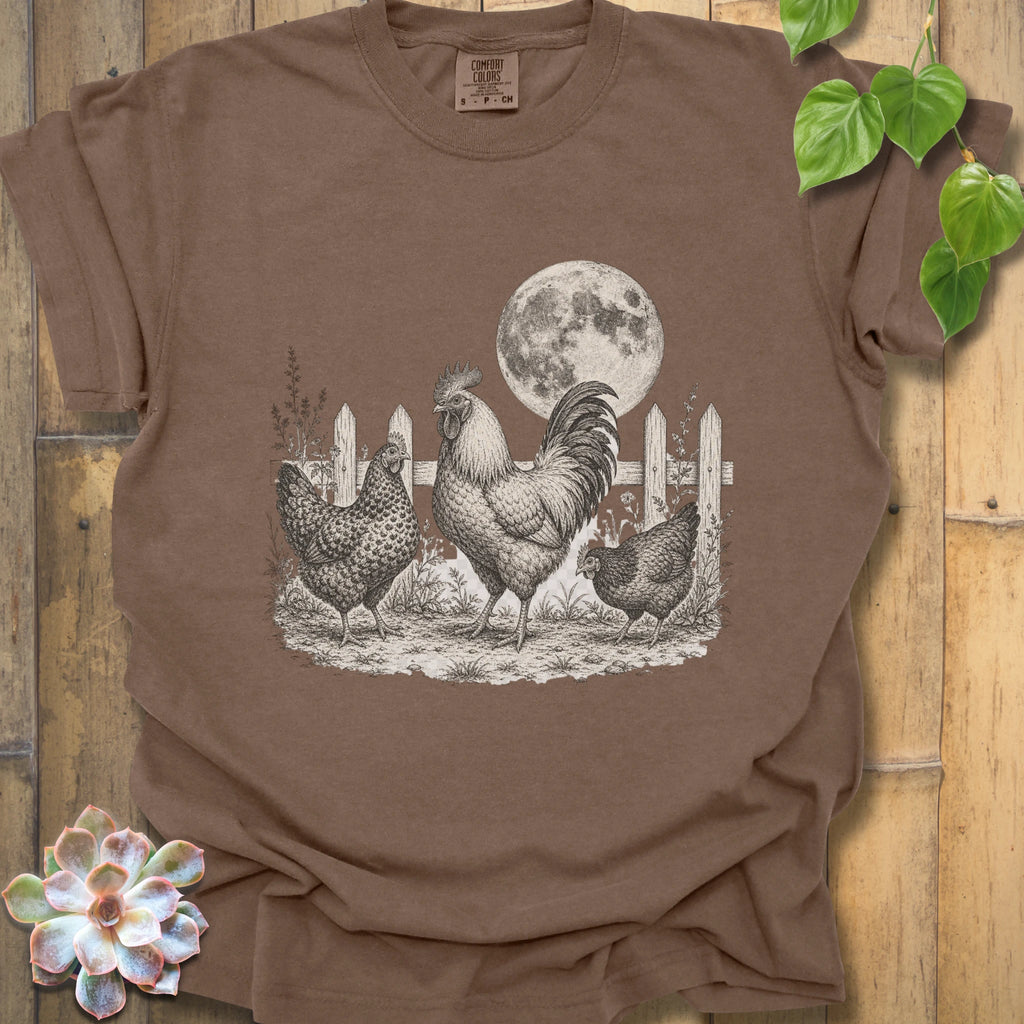 A brown short-sleeve crewneck t-shirt featuring a detailed black and white graphic of a rooster, two hens, a picket fence, and a full moon, laid flat on a rustic wooden background with decorative green leaves and a succulent.