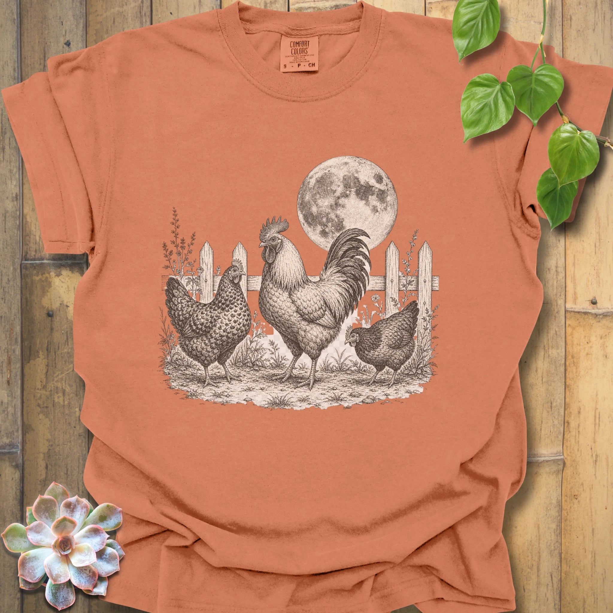 A terracotta orange short-sleeve t-shirt with a detailed black and white graphic of a rooster, two hens, a picket fence, and a full moon. The shirt is displayed in a flat-lay arrangement on a rustic wooden background, decorated with a succulent in the bottom-left corner and green leaves in the top-right.