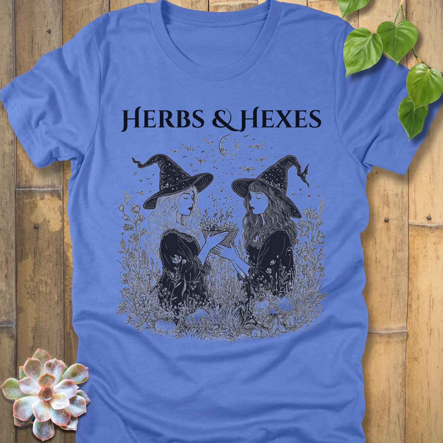 Heather Colombia Blue t-shirt featuring the design Herbs & Hexes in black letters, printed with two witches