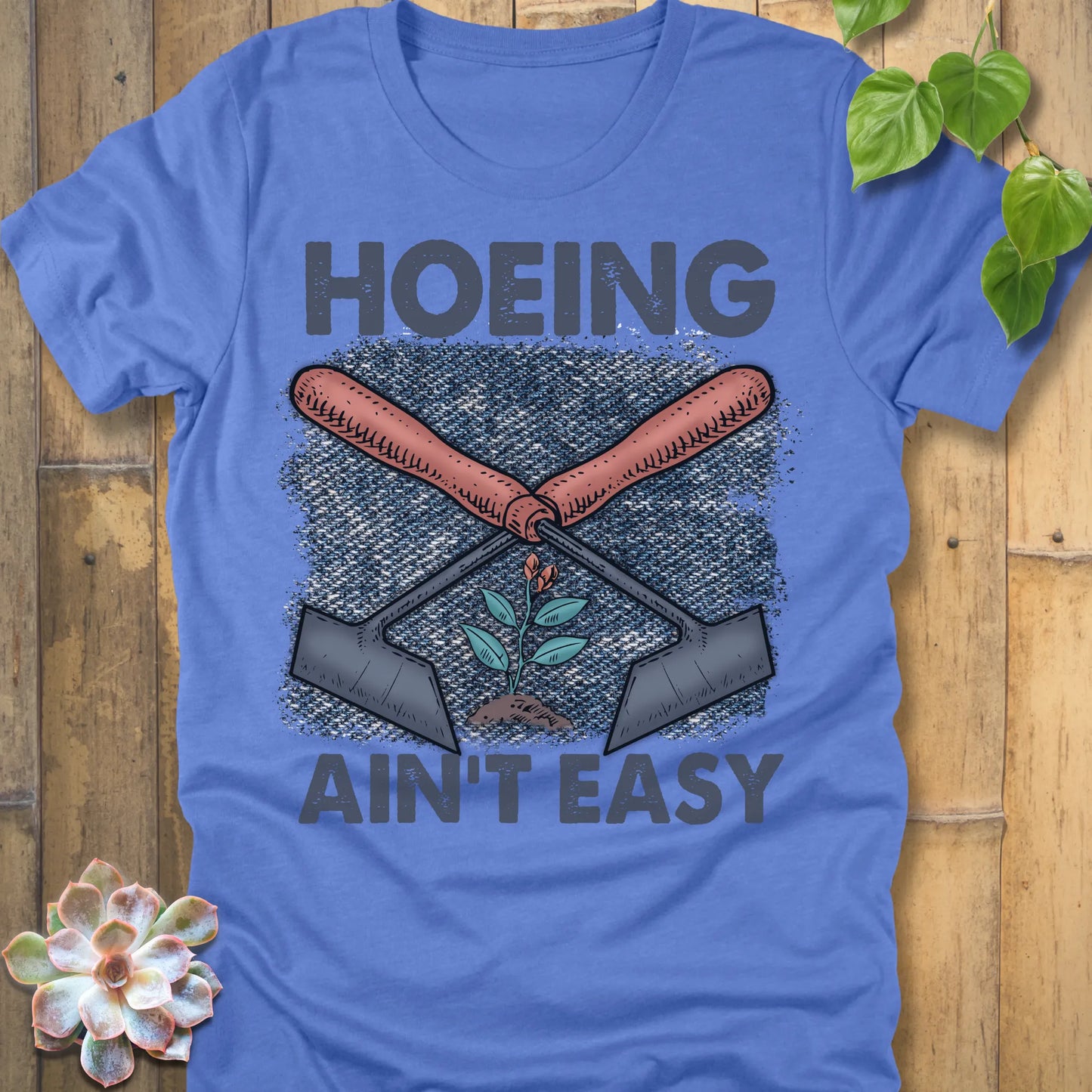Heather Columbia Blue t-shirt featuring the design Hoeing Ain’t Easy with gardening tools and plants printed in bold letters, funny gardening shirt.