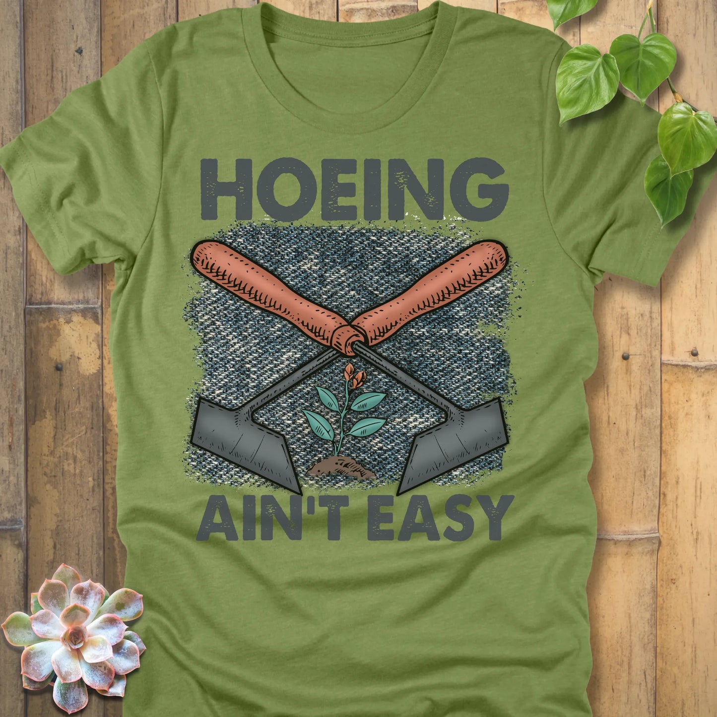 Heather Green t-shirt featuring the design Hoeing Ain’t Easy with gardening tools and plants printed in bold letters, funny gardening shirt.
