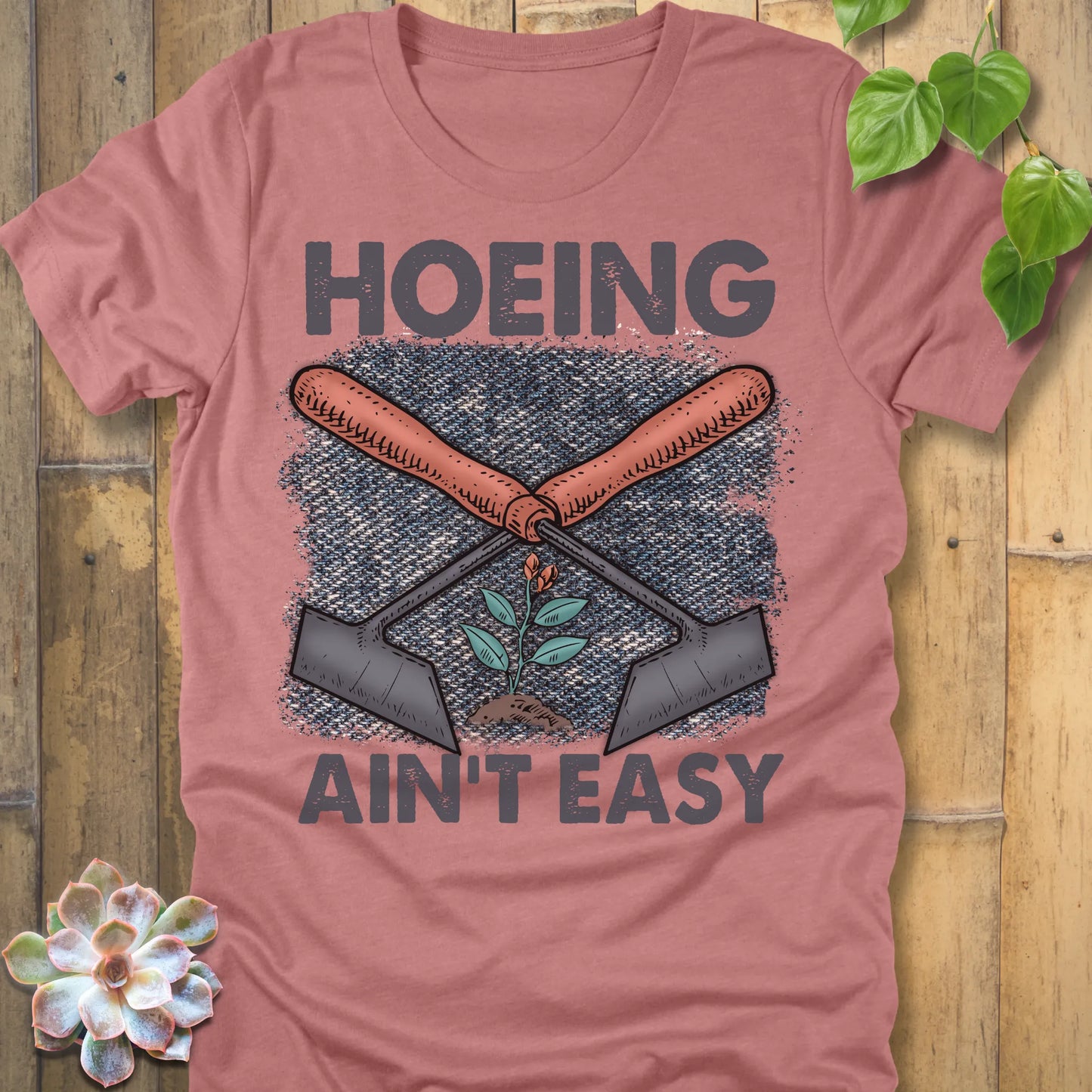 Heather Mauve t-shirt featuring the design Hoeing Ain’t Easy with gardening tools and plants printed in bold letters, funny gardening shirt.