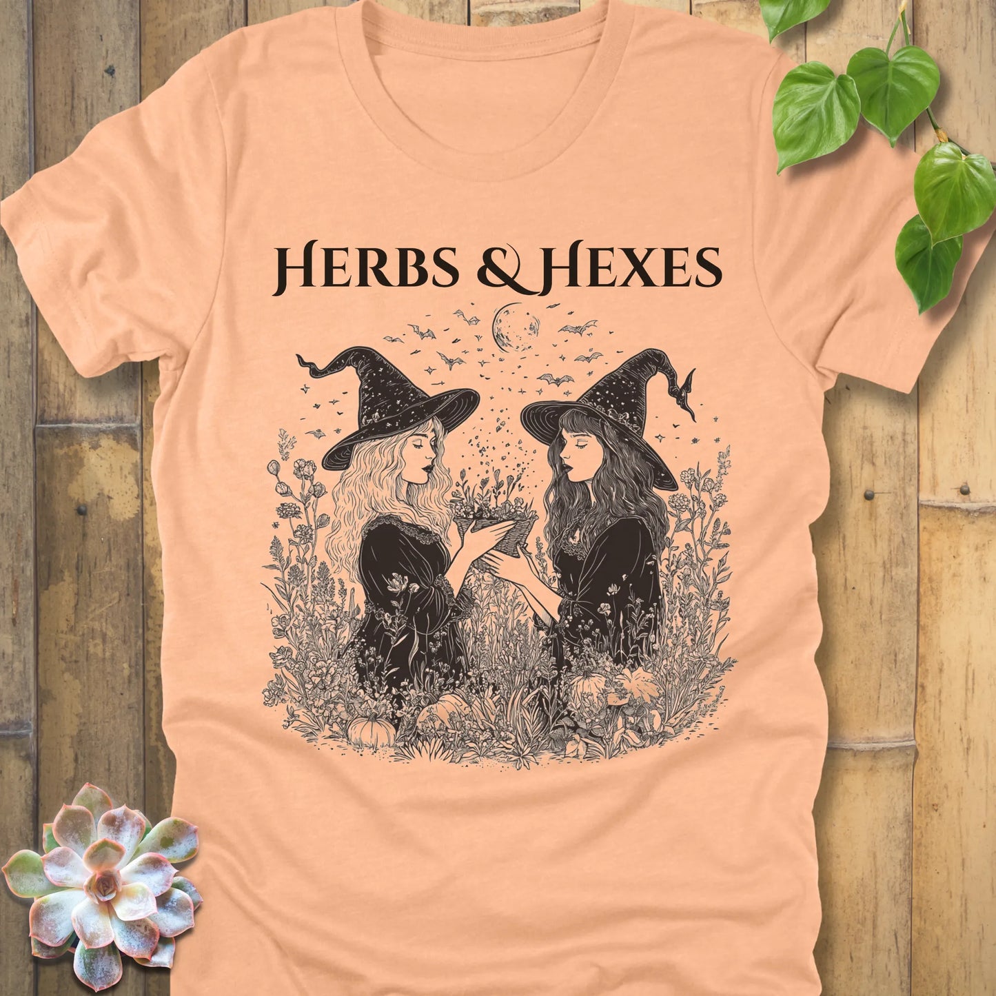Heather Peach t-shirt featuring the design Herbs & Hexes in black letters, printed with two witches