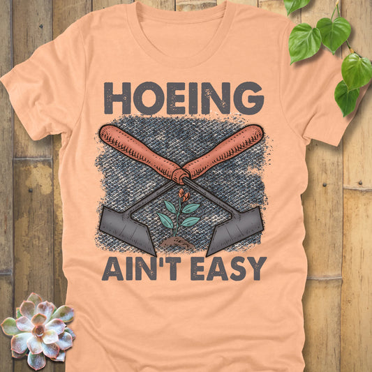 Heather Peach t-shirt featuring the design Hoeing Ain’t Easy with gardening tools and plants printed in bold letters, funny gardening shirt.