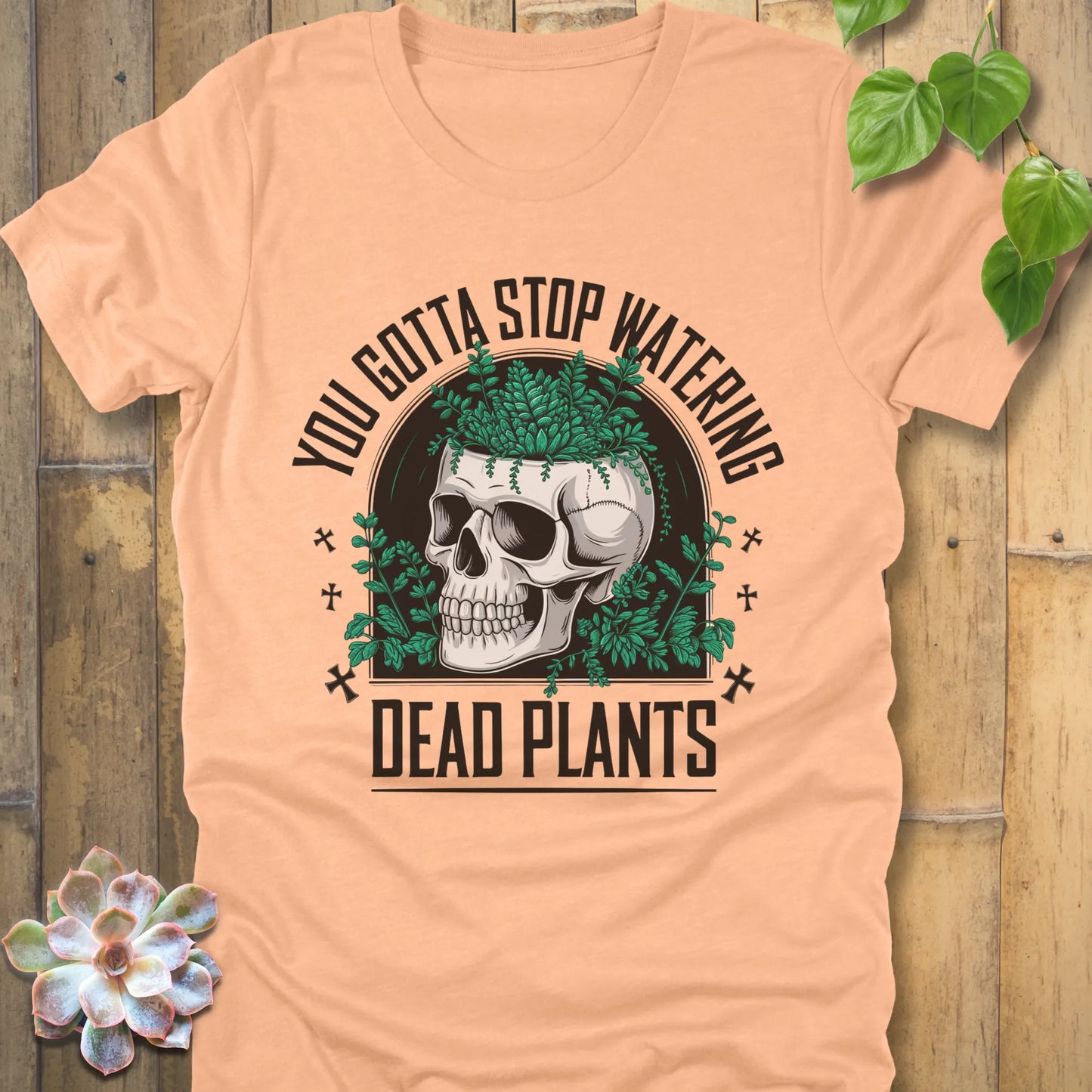 Heather Peach t-shirt featuring the design “You gotta stop watering dead plants” printed in black letters with skull and plants graphic
