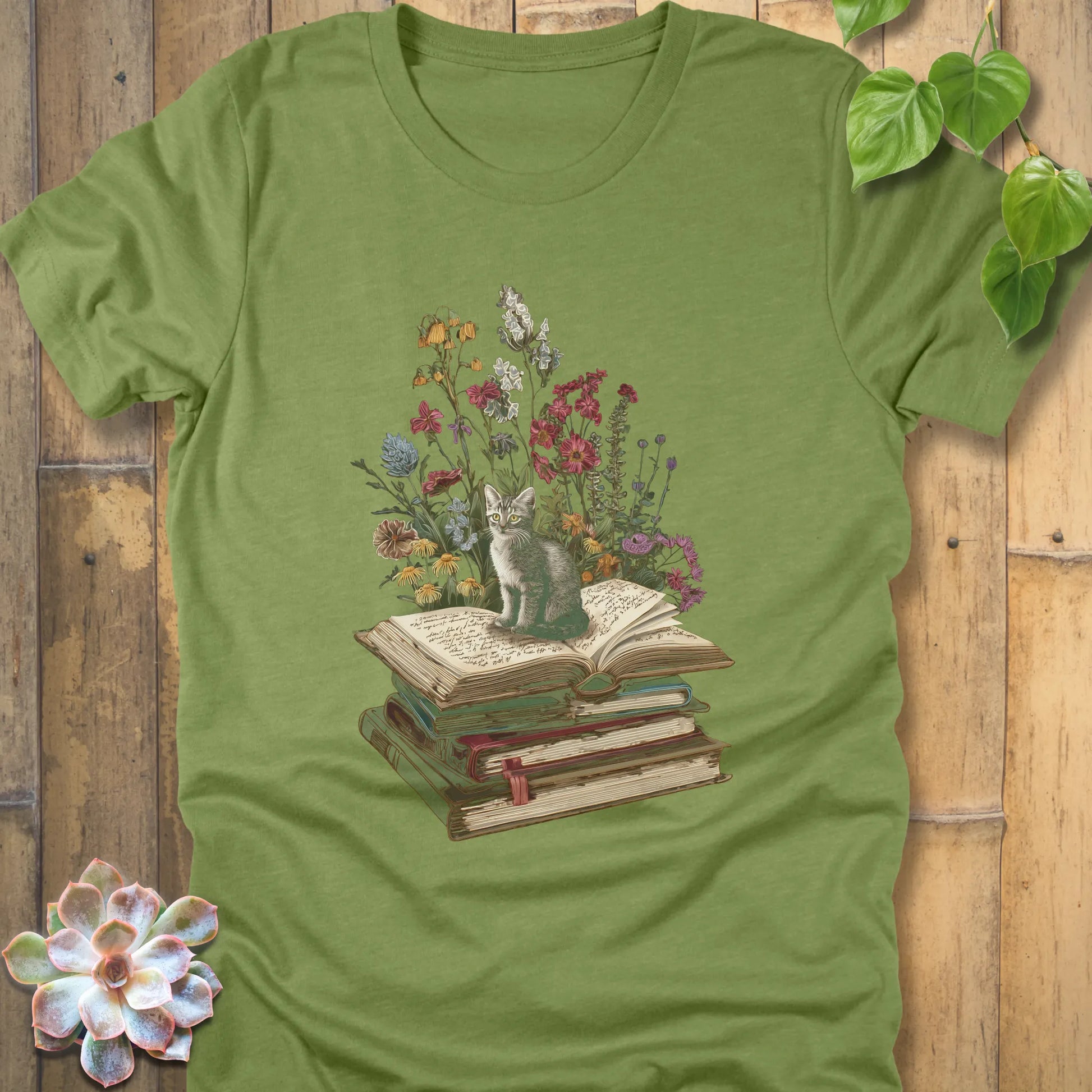 Heather green t-shirt featuring the design Whiskers & Words with a cat sitting on books and flowers, eco-style tee for book and cat lovers.