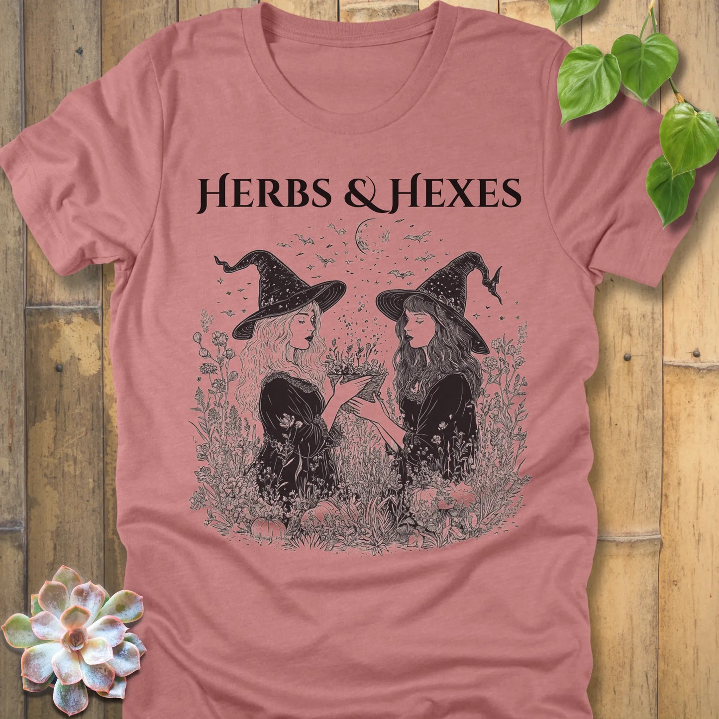 Heather mauve t-shirt featuring the design Herbs & Hexes in black letters, printed with two witches