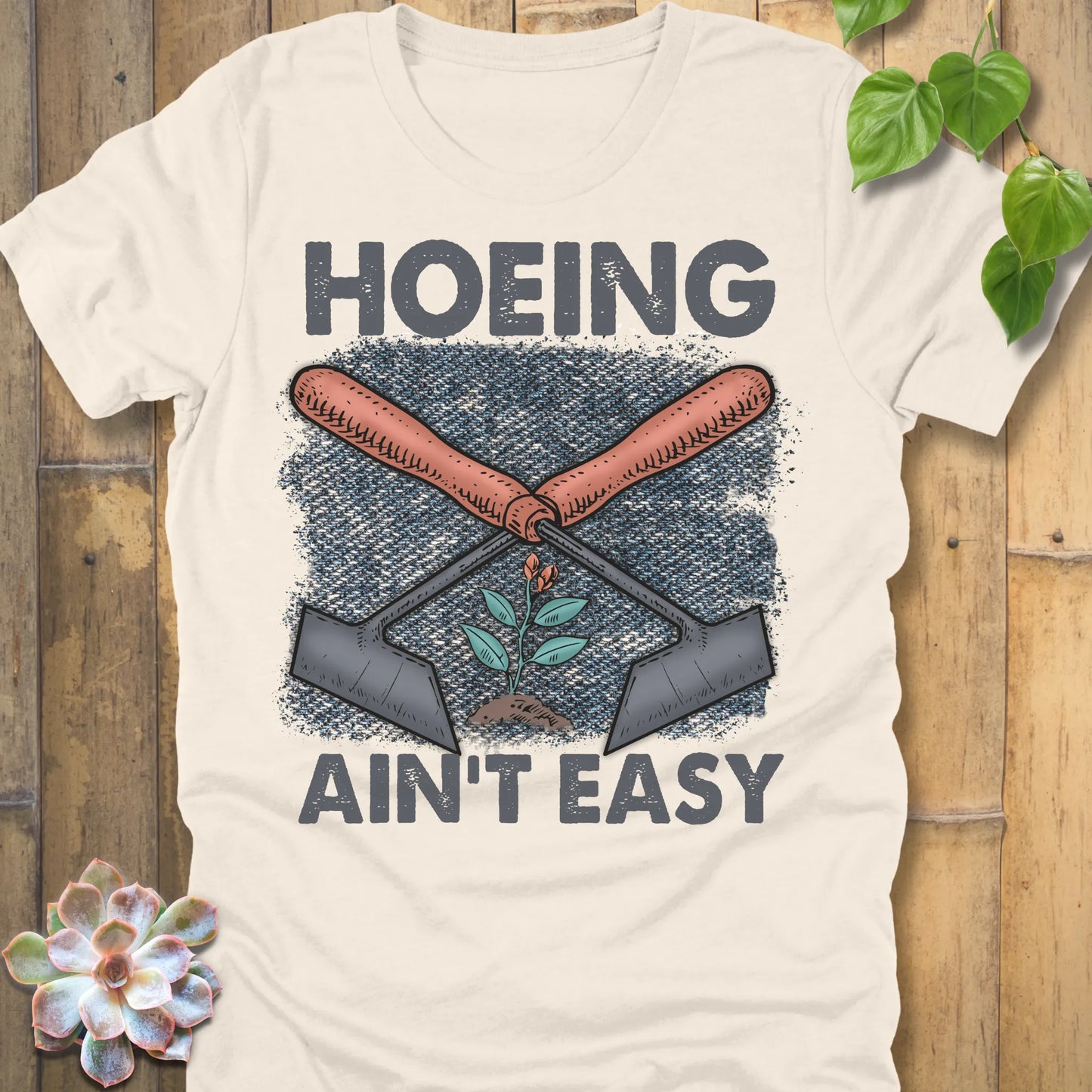 Natural t-shirt featuring the design Hoeing Ain’t Easy with gardening tools and plants printed in bold letters, funny gardening shirt.
